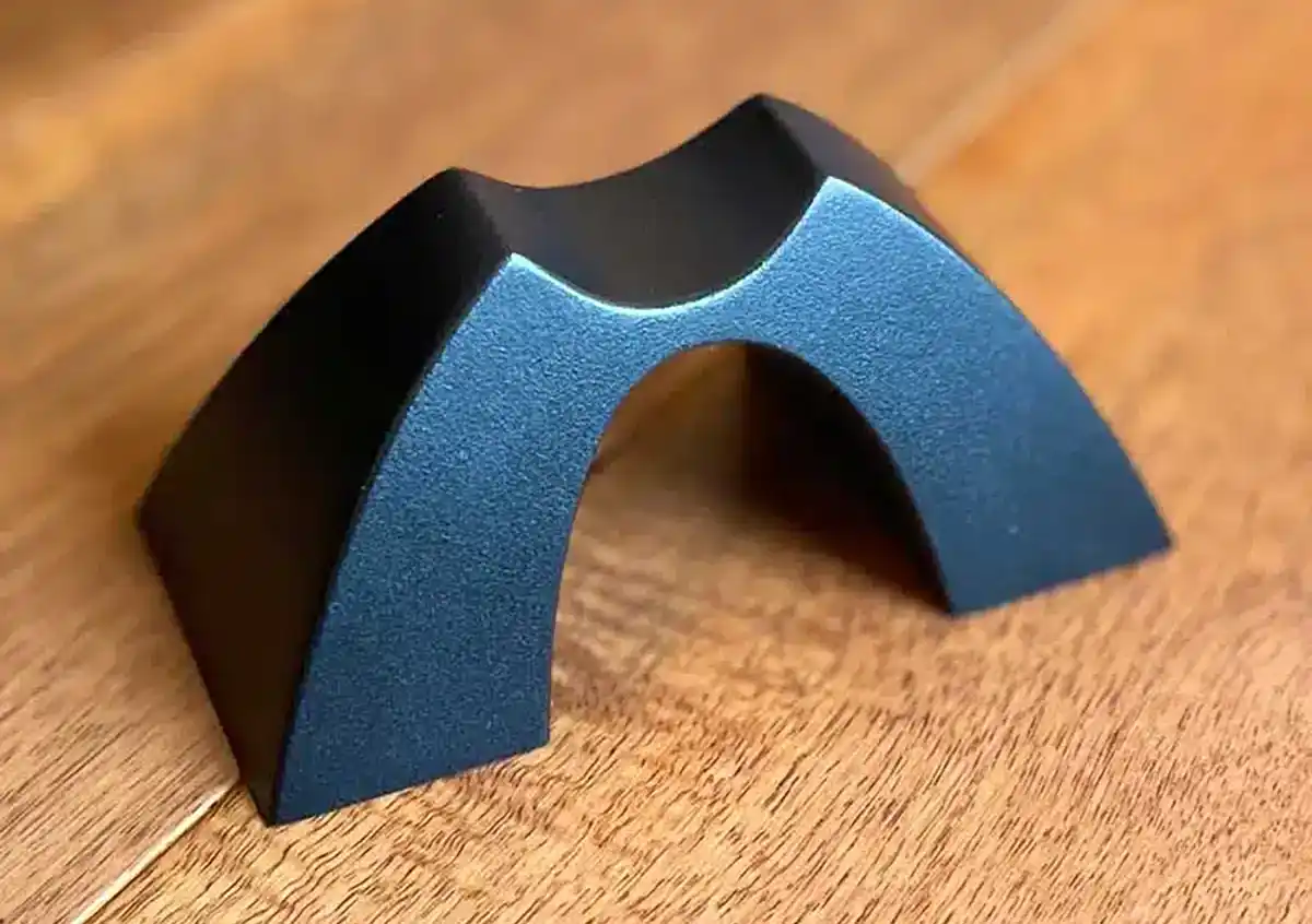Close-up product photo of a black Cable Arch speaker cable support with a curved top cradle and central arch opening on a wood floor.
